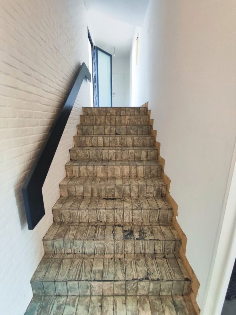 Escalier intérieur carrelé en cours de rénovation, avec mur blanc en briques et rampe noire